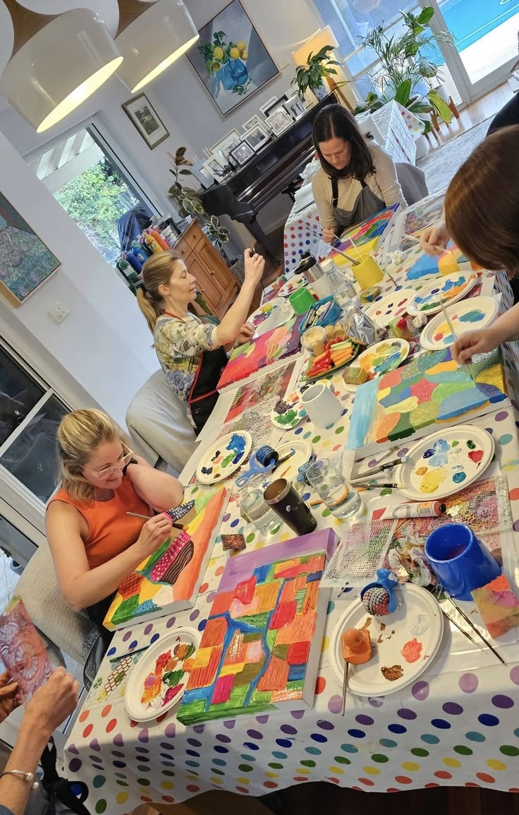 Pessoas pintando telas coloridas em volta de uma mesa com toalha de bolinhas